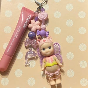 May include: A pink lip tint tube next to a keychain with a doll wearing a cat hat, a dolphin charm, and various pink and purple beads and charms. The keychain has a heart-shaped clasp.