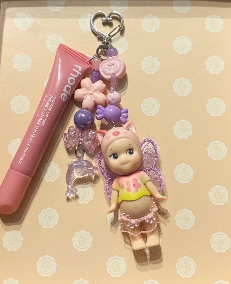 May include: A pink lip tint tube next to a keychain with a doll wearing a cat hat, a dolphin charm, and various pink and purple beads and charms. The keychain has a heart-shaped clasp.