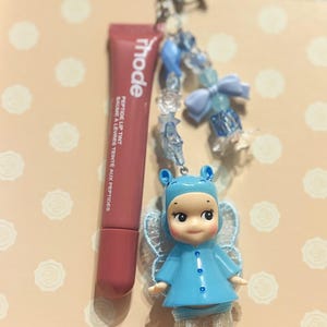 May include: A lip tint tube and a charm featuring a blue doll with orange boots and a beaded chain with a star-shaped clasp. The lip tint is a muted rose color, and the doll has a blue outfit and lace wings.