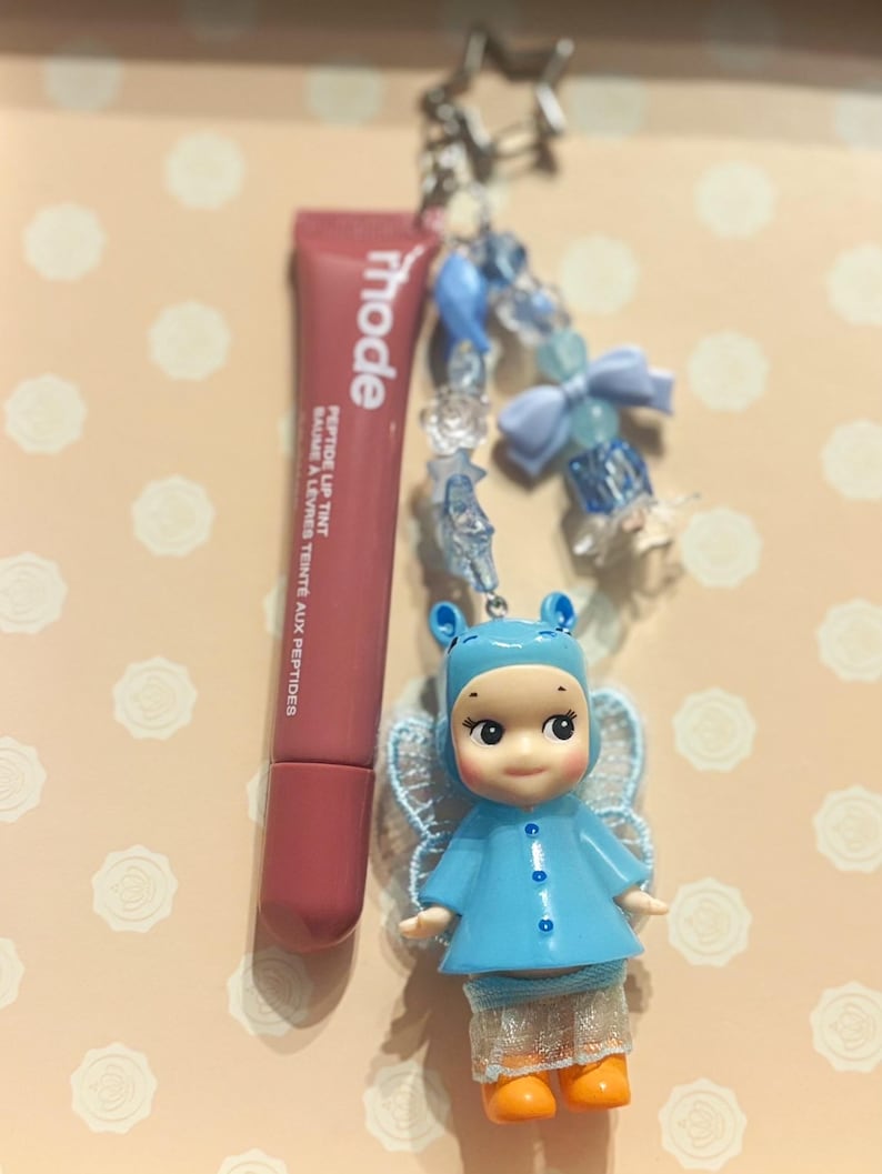 May include: A lip tint tube and a charm featuring a blue doll with orange boots and a beaded chain with a star-shaped clasp. The lip tint is a muted rose color, and the doll has a blue outfit and lace wings.