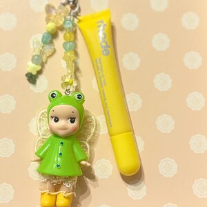 May include: A whimsical keychain featuring a small doll dressed in a green frog costume with yellow boots and a beaded butterfly charm. A yellow tube of lip tint with the word "rhode" is also present.