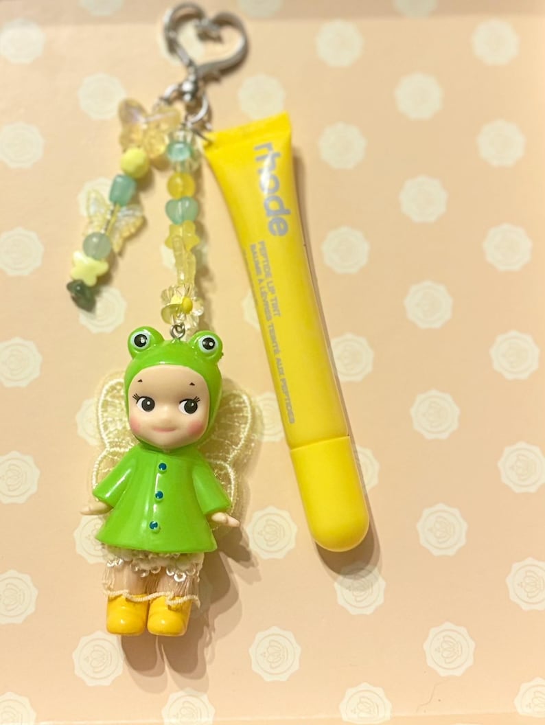 May include: A whimsical keychain featuring a small doll dressed in a green frog costume with yellow boots and a beaded butterfly charm. A yellow tube of lip tint with the word "rhode" is also present.