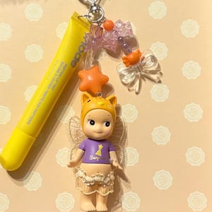 May include: A yellow lip tint tube and a charm featuring a small doll with a cat-eared hat, a purple shirt with a star graphic, and a skirt. The charm also includes orange star and bow accents, and a silver heart-shaped clasp.
