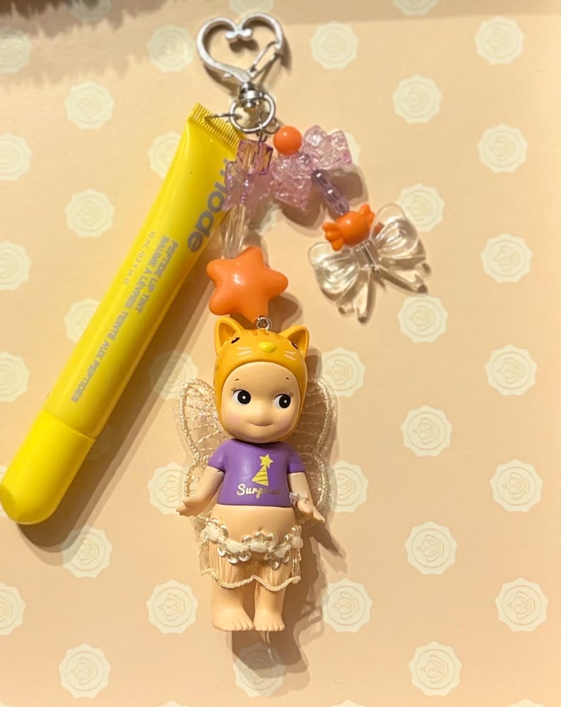 May include: A yellow lip tint tube and a charm featuring a small doll with a cat-eared hat, a purple shirt with a star graphic, and a skirt. The charm also includes orange star and bow accents, and a silver heart-shaped clasp.