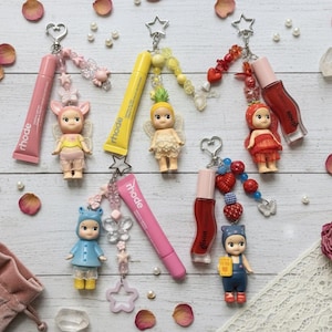May include: An assortment of keychains featuring small doll figures and lip gloss tubes. The dolls are dressed in various outfits and have wings. The lip gloss tubes are in pink and yellow, with the brand name "rhode" printed on them. Decorative elements include beads, charms, and rose petals.
