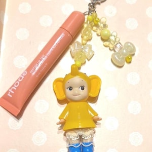 May include: A peach-colored lip tint tube with the brand name "rhode" next to a yellow elephant-themed charm. The charm has a yellow dress, blue boots, and lace shorts. A heart-shaped keychain and beaded accents complete the design.