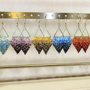 Op de afbeelding: Een display van kralen oorbellen in verschillende kleuren, waaronder wit, blauw, paars, goud, oranje, groen en zwart. Elke oorbel heeft een geometrisch ontwerp met een driehoekige bovenkant en een ruitvormige onderkant, hangend aan goudkleurige haken.