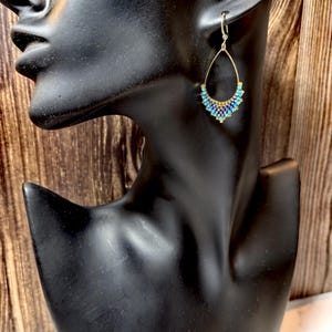 Può includere: Orecchini a goccia color oro con un design di perline. Gli orecchini presentano una montatura dorata con un motivo di perline nelle tonalità del blu, del viola e dell'oro. Gli orecchini sono esposti su una testa di manichino nera su sfondo di legno.