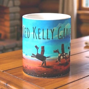 May include: A cylindrical ceramic mug with the text "NED KELLY GANG" in black. The mug features a landscape scene with stylized figures against a blue and red sky. The design is printed around the mug's exterior.