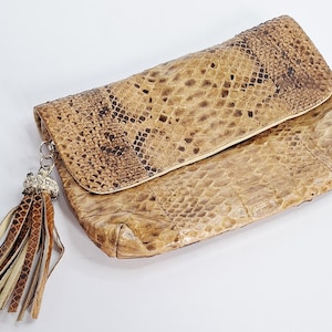 May include: A beige and brown snakeskin-patterned clutch with a fold-over flap. The clutch features a silver chain and a tassel with brown and beige leather strips. The bag is made of a textured material.