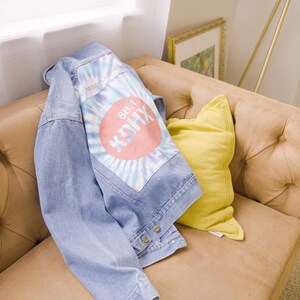Puede incluir: Chaqueta vaquera azul claro con un parche teñido anudado con el texto "88.1 KDHX" en un círculo rojo. La chaqueta está sobre un sofá color canela con mechones junto a un cojín amarillo.