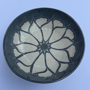 May include: A ceramic bowl with a dark blue exterior and a cream-colored floral design. The flower pattern is centered in the bowl and has a detailed, hand-painted appearance. The bowl's rim is also dark blue.