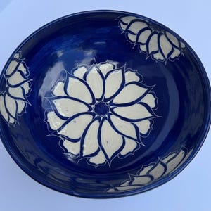 May include: A deep blue ceramic bowl with a glossy finish. The bowl features a large, white floral design in the center, surrounded by smaller floral patterns around the rim. The design is hand-painted.