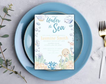 May include: A baby shower invitation with an "Under the Sea" theme, featuring aquatic illustrations and the text "Join Us Under the Sea". The invitation is on a light blue plate, with gold cutlery and a sprig of greenery.