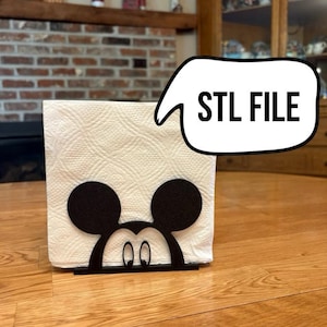 Puede incluir: Un portapañuelos de metal negro con el tema de Mickey Mouse. El soporte presenta un diseño recortado de la cara y las orejas de Mickey, sosteniendo una pila de toallas de papel blancas. El texto "STL FILE" está en un bocadillo.