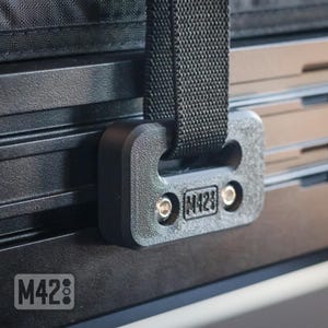 May include: A close-up shot of a black latching mechanism with a woven strap. The mechanism has the text "M42" and "M428" printed on it. The background is a textured, black surface.