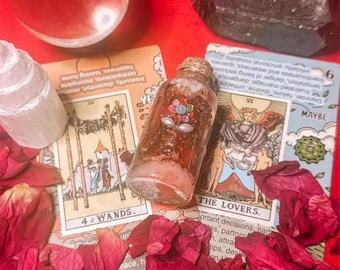 Op de afbeelding: Een heldere glazen fles met een decoratieve stop, gevuld met een roodachtige vloeistof en versierd met kleurrijke versieringen, ligt tussen tarotkaarten en rode rozenblaadjes. Een kristallen bol en een zwarte kristal zijn ook aanwezig.