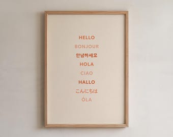 May include: A rectangular wall art print in a light wood frame featuring the word hello in eight different languages stacked vertically in alternating shades of orange and light pink against a cream-colored background.
