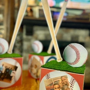 May include: Baseball-themed decorative boxes with a baseball and a wooden bat. The boxes have a red and white baseball design with a photo of a family. The boxes are topped with artificial green grass.