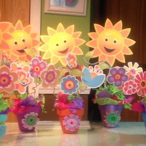 May include: Five colorful flower centerpieces with smiling yellow suns, green grass, and colorful flowers in pots. The pots are decorated with colorful flowers and butterflies.