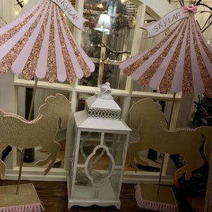May include: Two gold glitter horse centerpieces with pink glitter umbrellas and the name "Skylar" on each. The umbrellas are on gold sticks and the horses are on gold stands with pink fringe. A white lantern is in the center.