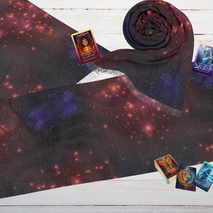 Pode incluir: Um lenço preto com estampa de galáxia, apresentando nebulosas e estrelas vermelhas e roxas. O lenço tem franjas nas extremidades e é acompanhado por vários cartões de tarô com ilustrações coloridas. Em uma superfície de madeira branca.