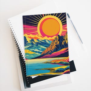 May include: A spiral-bound notebook featuring a vibrant landscape illustration. The cover showcases a large orange sun with radiating lines, mountains, and a lake in shades of blue, yellow, and pink. A pen rests beside the notebook.