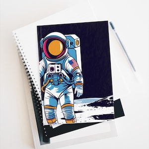 May include: A spiral-bound notebook with a cover featuring a retro illustration of an astronaut on a moon-like surface. The astronaut is wearing a white and blue spacesuit with an American flag patch. A pen rests on the white background.