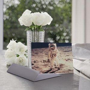 May include: A display of white roses in a clear glass vase, a greeting card depicting an astronaut on the moon, and a grey card holder. The background shows a window with heart-shaped bokeh.