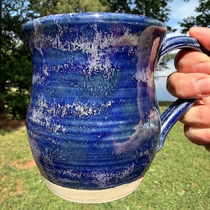 May include: A handmade ceramic mug with a deep blue glaze and a textured, speckled surface. The mug has a rounded body, a sturdy handle, and a light beige base. The mug is designed for hot beverages.