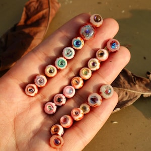 Puede incluir: Una mano que sostiene varias filas de pequeñas cuentas de ojo de vidrio de colores. Las cuentas tienen una variedad de colores de iris, incluyendo azul, verde y rosa, con detalles rojos y blancos en los bordes. El fondo es borroso y de tono neutro.