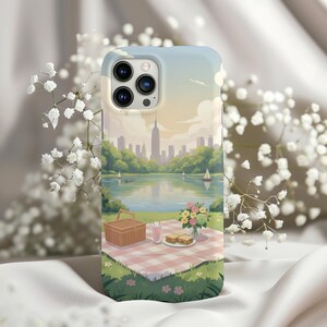 May include: A phone case featuring a scenic illustration of a picnic in a park with a lake and city skyline. The case has a light blue sky background with a picnic basket, food, and flowers on a checkered blanket.