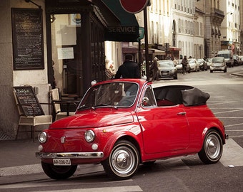 Op de afbeelding: Een felrode vintage Fiat 500 cabriolet geparkeerd op een straat in de stad. De auto heeft een zwart canvas dak en een chromen bumper. De auto staat geparkeerd voor een gebouw met een bord met de tekst "Service".