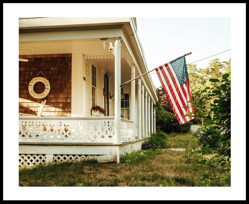 May include: A white wooden house with a porch and a white picket fence. An American flag hangs from the porch, waving in the breeze. The house has a wooden sign that reads "North Star New York".