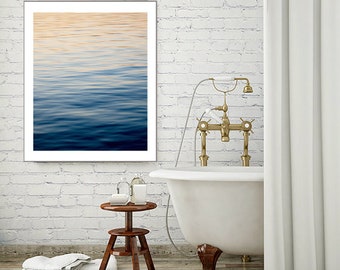 May include: A framed photograph of a blue and white abstract water scene hangs above a white bathtub with gold fixtures. The bathroom has white brick walls and a white shower curtain.