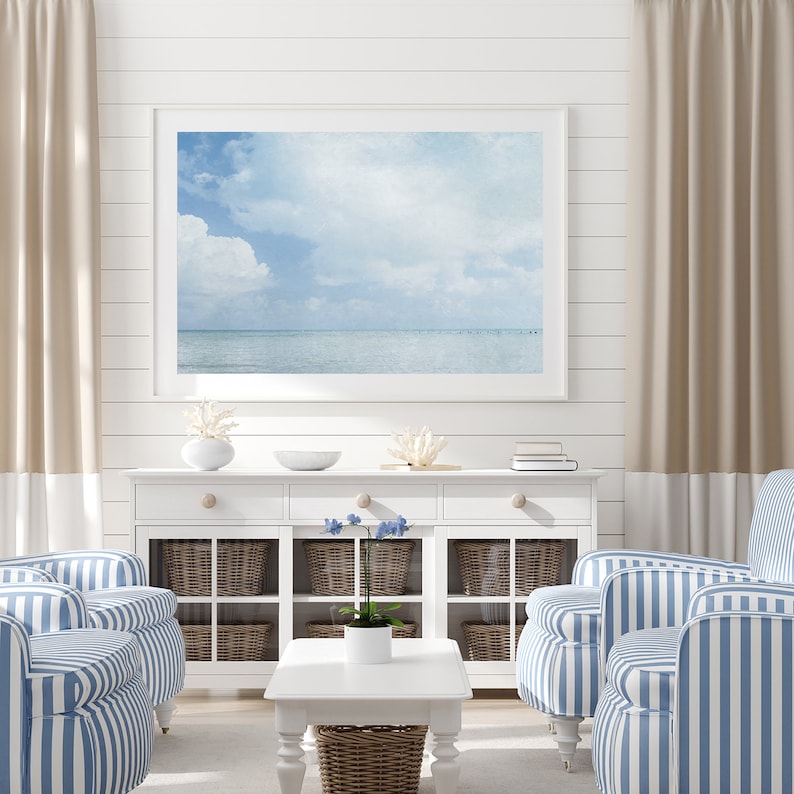 May include: A white living room with two blue and white striped armchairs, a white coffee table, a white cabinet with glass doors and wicker baskets, and a framed photograph of a seascape with a blue sky and white clouds.