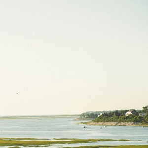 Może przedstawiać: Widok na spokojne zbiornik wodny z trawiastym brzegiem i kilkoma domami w oddali. Niebo jest jasnoniebieskie, a woda ma jasnoniebiesko-zielony kolor.