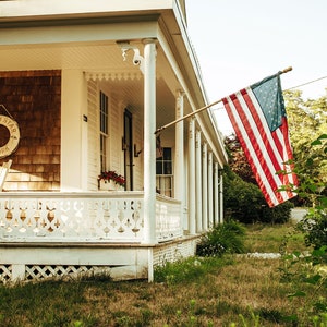 May include: A white house with a porch and a wooden railing. An American flag is flying from a pole on the porch. The house has a brown wooden siding and a white trim. There is a life preserver hanging on the wall with the words "North Star New York" on it.