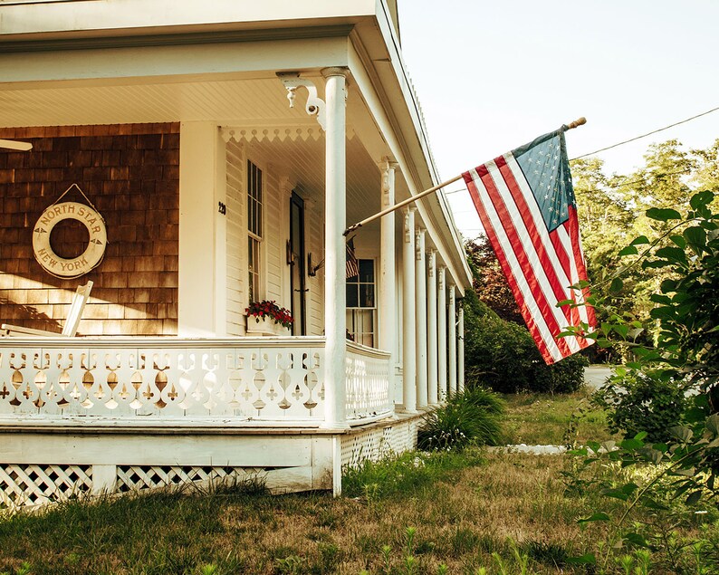 May include: A white house with a porch and a wooden railing. An American flag is flying from a pole on the porch. The house has a brown wooden siding and a white trim. There is a life preserver hanging on the wall with the words "North Star New York" on it.