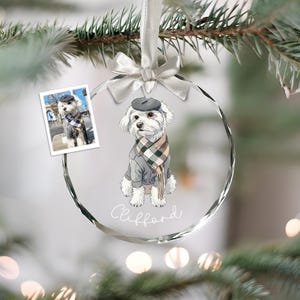 Op de afbeelding: Een heldere, ronde kerstbal met een aquarel illustratie van een hond met een baret, trui en sjaal. De naam van de hond, "Clifford", staat eronder geschreven. Een kleine foto van de hond is aan de ornament bevestigd.