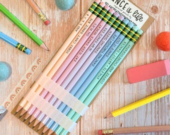 May include: A set of ten pastel-colored pencils arranged on a cardboard backing labeled DANCE IS LIFE. Each pencil features a unique dance-themed phrase including AND 5, 6, 7, 8, SORRY, CAN'T. DANCE. BYE., OH. PLIES..., EAT. SLEEP. DANCE. REPEAT., IN MY DANCE ERA, DANCING QUEEN, DANCING THROUGH LIFE, DANCE IT OUT, ATHLETE + ARTIST = DANCER, and COFFEE AND CHOREOGRAPHY.