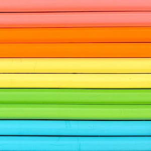 May include: A series of parallel wooden pencils arranged horizontally in a spectrum of colors. The sequence features rows of coral, orange, yellow, lime green, and light blue pencils, each with a smooth, matte finish and distinct dark lines.