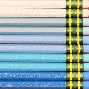 May include: A collection of ten wooden pencils arranged in a gradient from white to dark blue. Each pencil features a speckled finish, a metallic green ferrule with two yellow stripes, and a light blue eraser at the end of the writing instrument.