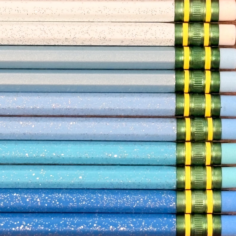 May include: A collection of ten wooden pencils arranged in a gradient from white to dark blue. Each pencil features a speckled finish, a metallic green ferrule with two yellow stripes, and a light blue eraser at the end of the writing instrument.
