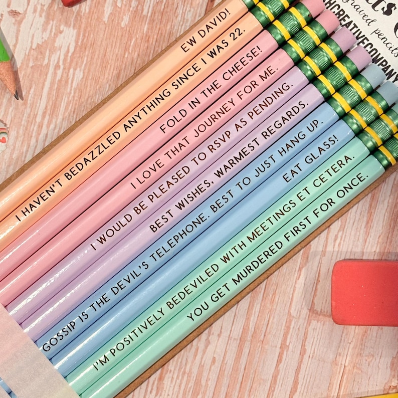 May include: A set of ten pastel-colored wooden pencils arranged in a row, each featuring a unique quote printed in black text. The pencils have metallic gold ferrules with green erasers and include phrases like EW DAVID, FOLD IN THE CHEESE, and EAT GLASS.