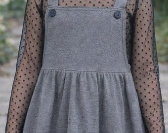 May include: A young girl wearing a gray jumper dress with black long sleeves. The dress has a flared skirt and button details on the straps. The black sleeves have a polka dot pattern.