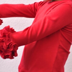 Può includere: Maglietta rossa a maniche lunghe con polsini a volant. La maglietta è realizzata in un tessuto morbido ed elastico.