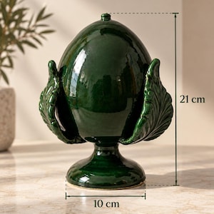 Può includere: Un ornamento in ceramica verde scuro lucido a forma di pigna su base a piedistallo, caratterizzato da scaglie scolpite a forma di foglia. L'oggetto misura 21 cm in altezza e 10 cm in larghezza, come indicato dalle linee di misura e dal testo.