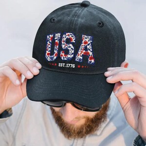 Puede incluir: Gorra de b&eacute;isbol negra con la palabra "USA" en un dise&ntilde;o floral rojo, blanco y azul. El texto "EST. 1776" est&aacute; debajo. La gorra tiene una visera curva y un bot&oacute;n en la parte superior.