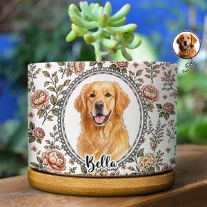 Puede incluir: Una maceta cilíndrica de cerámica con un patrón floral y un marco ovalado central que contiene una ilustración de un golden retriever con el nombre Bella escrito debajo. La maceta se asienta sobre una base de madera y contiene una suculenta.